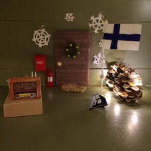 Tonttuoven edessä pöytä, jolla on vanhanaikainen radio. Oven oikealla puolen lipputanko, jossa on Suomen lippu. Pieni partiohuivi lipputangon juurella.