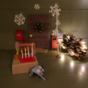 Tonttuoven edustalla pieni pöytä, jonka päällä on radio, hyasintti ja adventtikynttelikkö. Neljässä kynttilässä liekki. Pöydän edessä pieni partiohuivi.